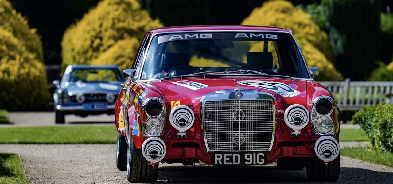 Two classic Mercedes-Benz cars on a country driveway — a red AMG-liveried saloon in front and a blue vintage Mercedes behind